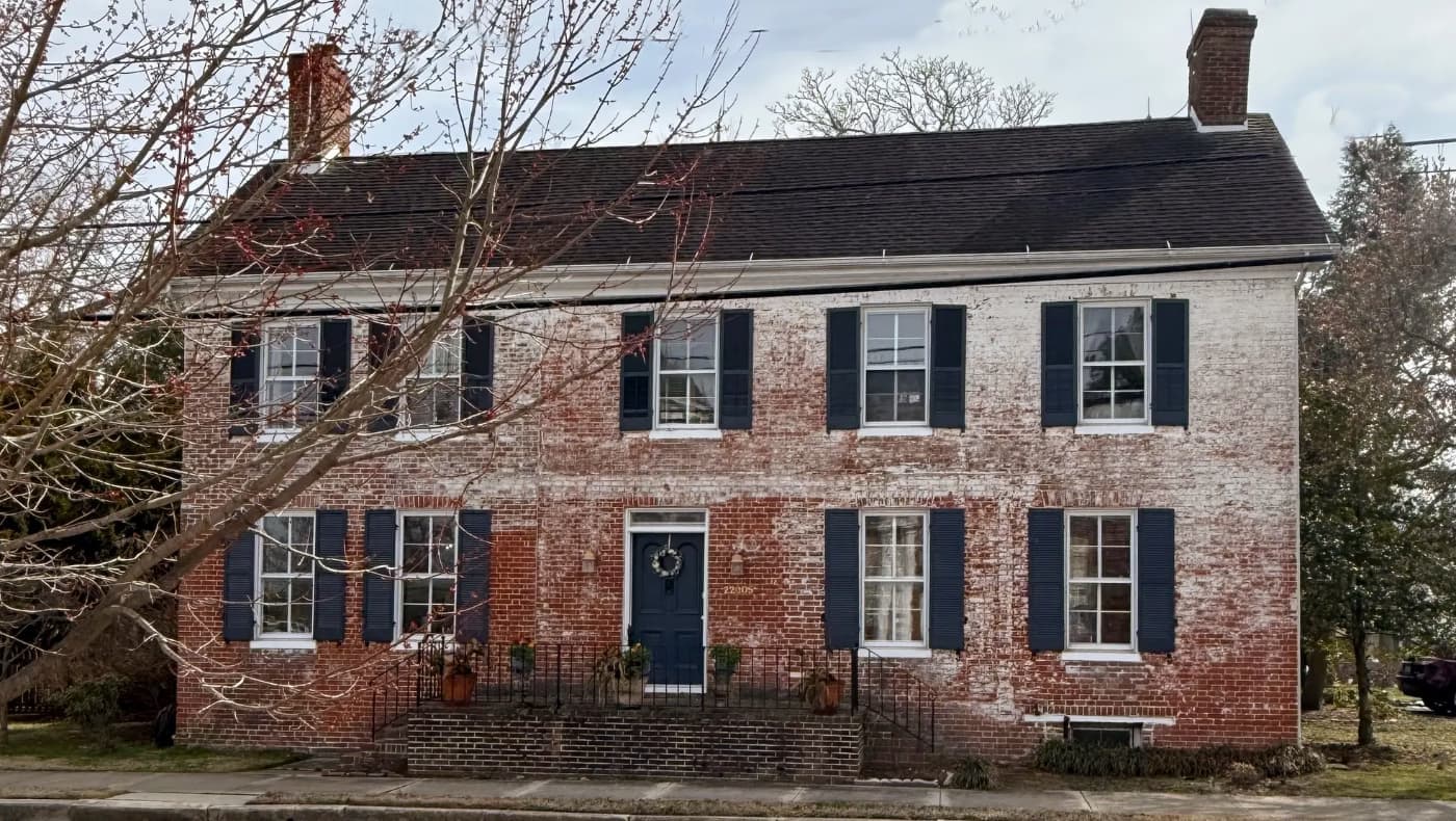 A stately two-story red brick Colonial home, built in 1794, featuring a symmetrical facade, white trim, and mature shade trees on a level corner lot in Hillsboro, MD.