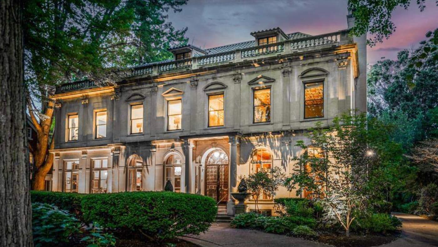 Front exterior of the Louis Kamper Mansion in Indian Village, Detroit. This 1915 Neo-Renaissance estate features a grand limestone facade, a symmetrical entry with a copper-roofed portico, and elegant arched windows surrounded by professional landscaping and wrought iron fencing.