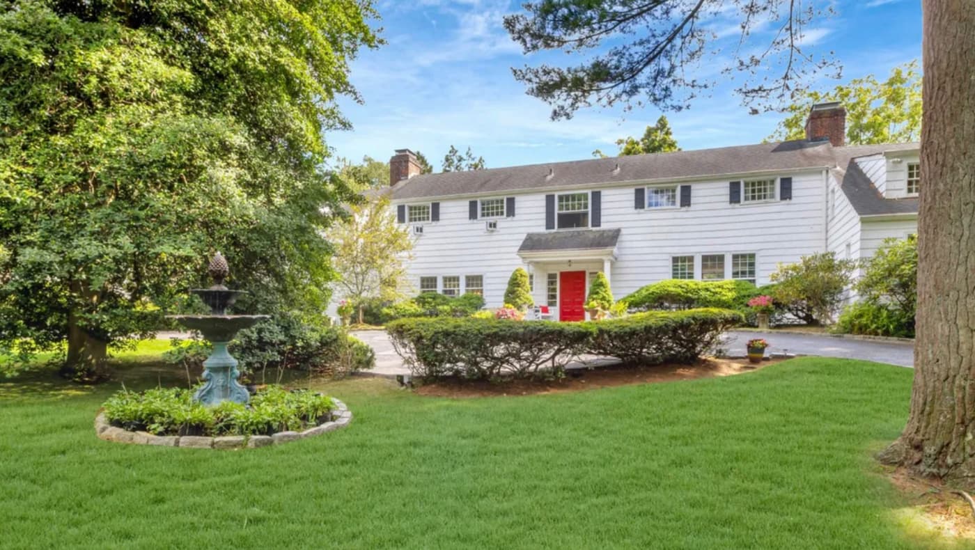 Exterior view of "Ben Robyn Farm," a c. 1910 Colonial Revival estate in Huntington, NY. The 5,020-square-foot white clapboard home features a symmetrical facade, a classic gabled roof, and multiple chimneys.