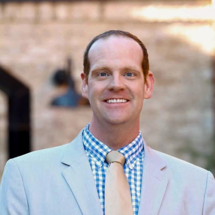 Professional headshot of Chess Thigpen, a licensed Georgia Realtor®, smiling in a light gray suit and blue checkered shirt, set against a blurred background of a historic stone building.