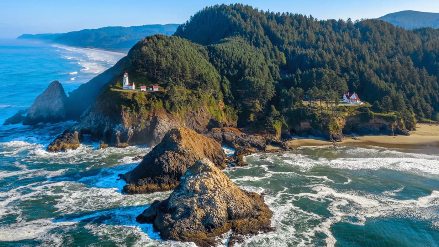 A panoramic seaward view of the Heceta Head Lighthouse and Lightkeeper’s House perched on a rugged emerald cliff along the Oregon Coast. The iconic white conical lighthouse tower stands tall on the rocky promontory, while the historic two-story Queen Anne-style keeper’s cottage sits nestled on the hillside below.
