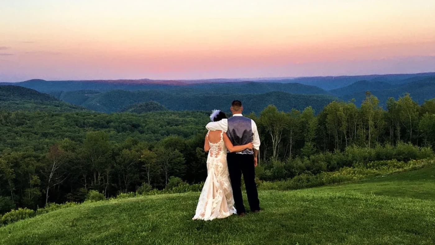 A lifestyle shot of a married couple standing on the elevated balcony of the Blue Vista Motor Lodge in North Adams, MA. They are looking out over a panoramic view of the Berkshire Mountains and the Hoosac Range at sunset.