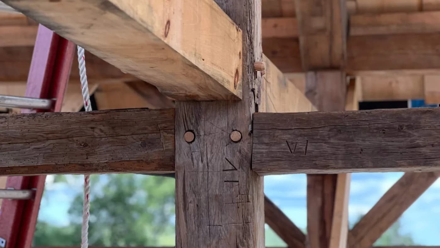 A detailed close-up of a structural restoration by Preservation Timber Framing, Inc. The image shows a skilled carpenter fitting a new, hand-planed white oak timber into an original 18th-century frame.
