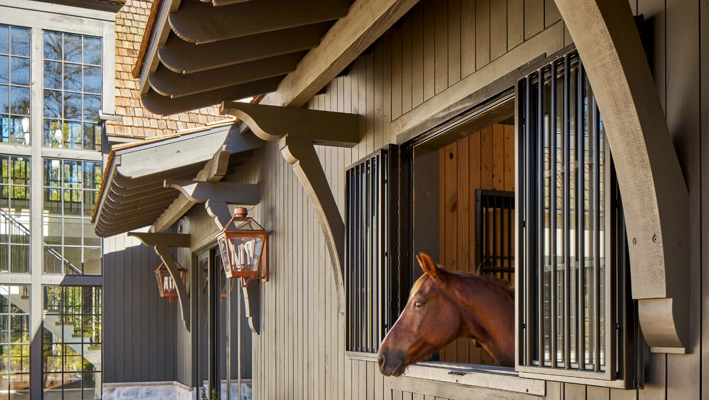 Exterior view of luxury horse stables featuring a striking timber-frame design by Frazier Home Design and Midtown Custom Homes. The structure showcases heavy, dark-stained timber gables and decorative trusses provided by Tar River Timber Works, contrasting beautifully against white vertical board-and-batten siding.