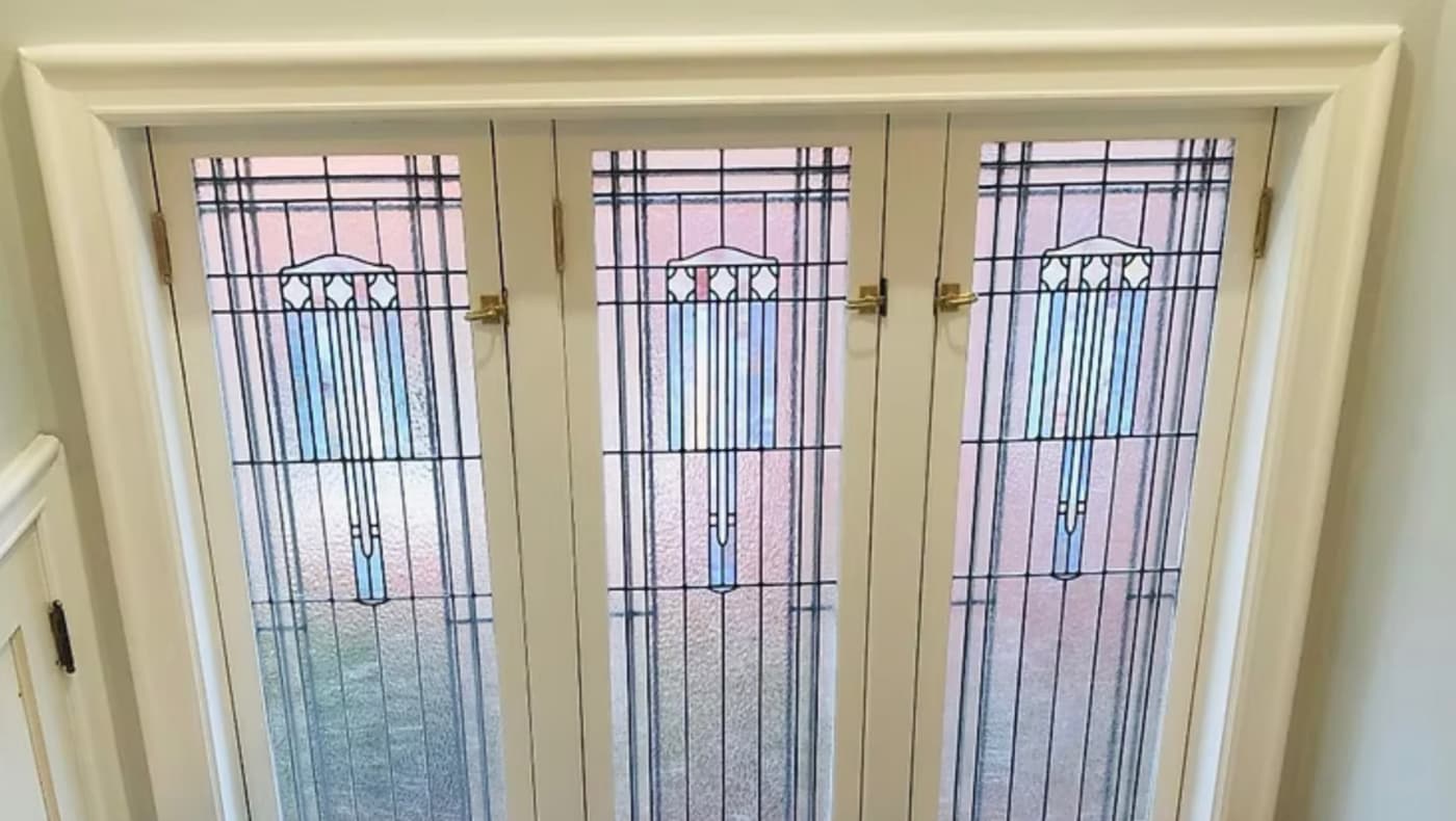 Three leaded-glass casement windows with white wood frames. The glass has a geometric Prairie School-style design, with vertical lines and a central decorative motif.