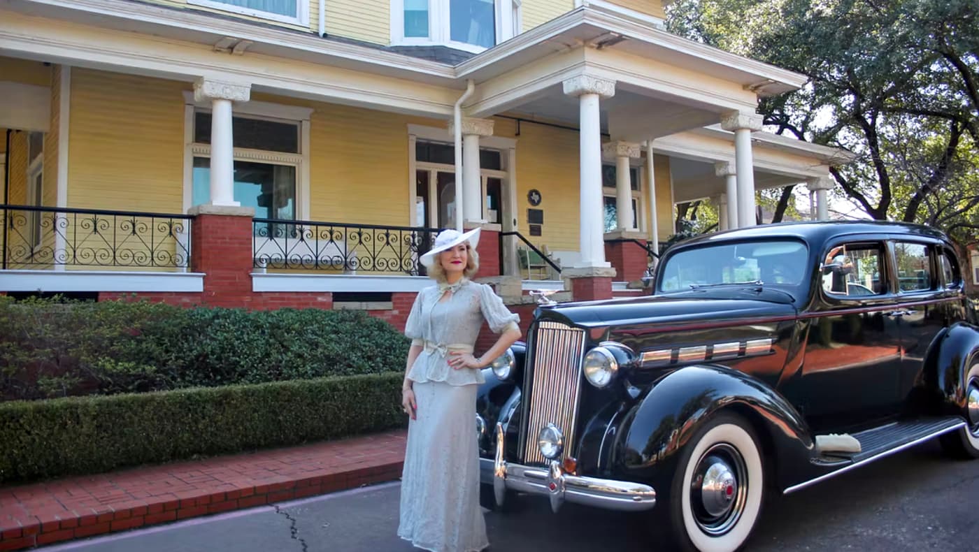 A vibrant exterior view of the historic 1900 Victorian-style Heard-Craig Center for the Arts. A woman in an elegant, vintage-inspired gown and wide-brimmed hat stands beside a polished classic luxury car parked on the driveway, recreating the Gilded Age sophistication of McKinney’s historic district.