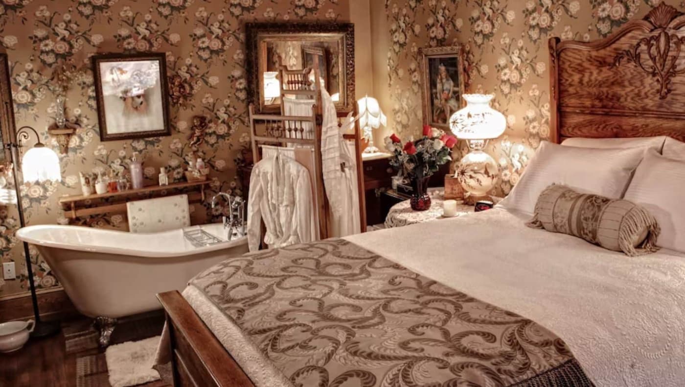 A lavishly decorated Victorian bedroom at The Queen Bed & Breakfast in Bellefonte, PA. The room features a grand antique carved wood bed frame with high-thread-count linens, surrounded by period-correct floral wallpaper and ornate window drapery.