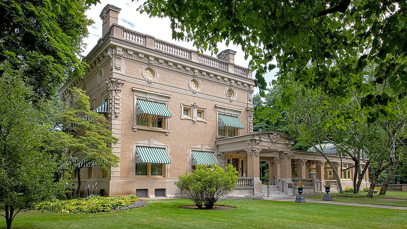 The front exterior of Ruthmere Mansion in Elkhart, Indiana. A three-story Beaux-Arts limestone and buff-brick residence featuring a grand symmetrical facade, a balustraded piazza, and a prominent porte-cochère.