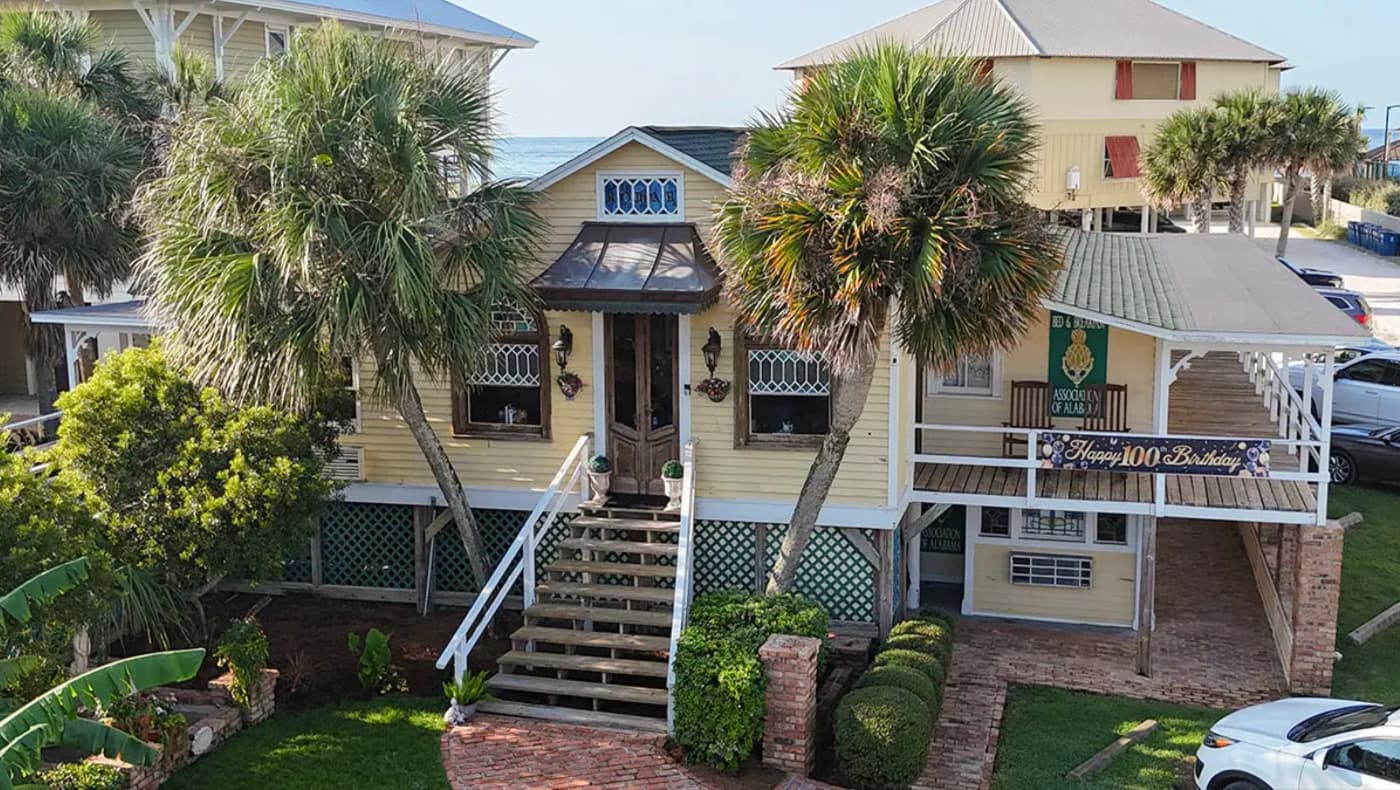 Exterior view of The Original Romar House Bed & Breakfast Inn in Orange Beach, Alabama. This historic c. 1924 beach cottage features a classic cypress-sided facade, a steep gabled roof, and a wide screened-in porch facing the Gulf of Mexico.