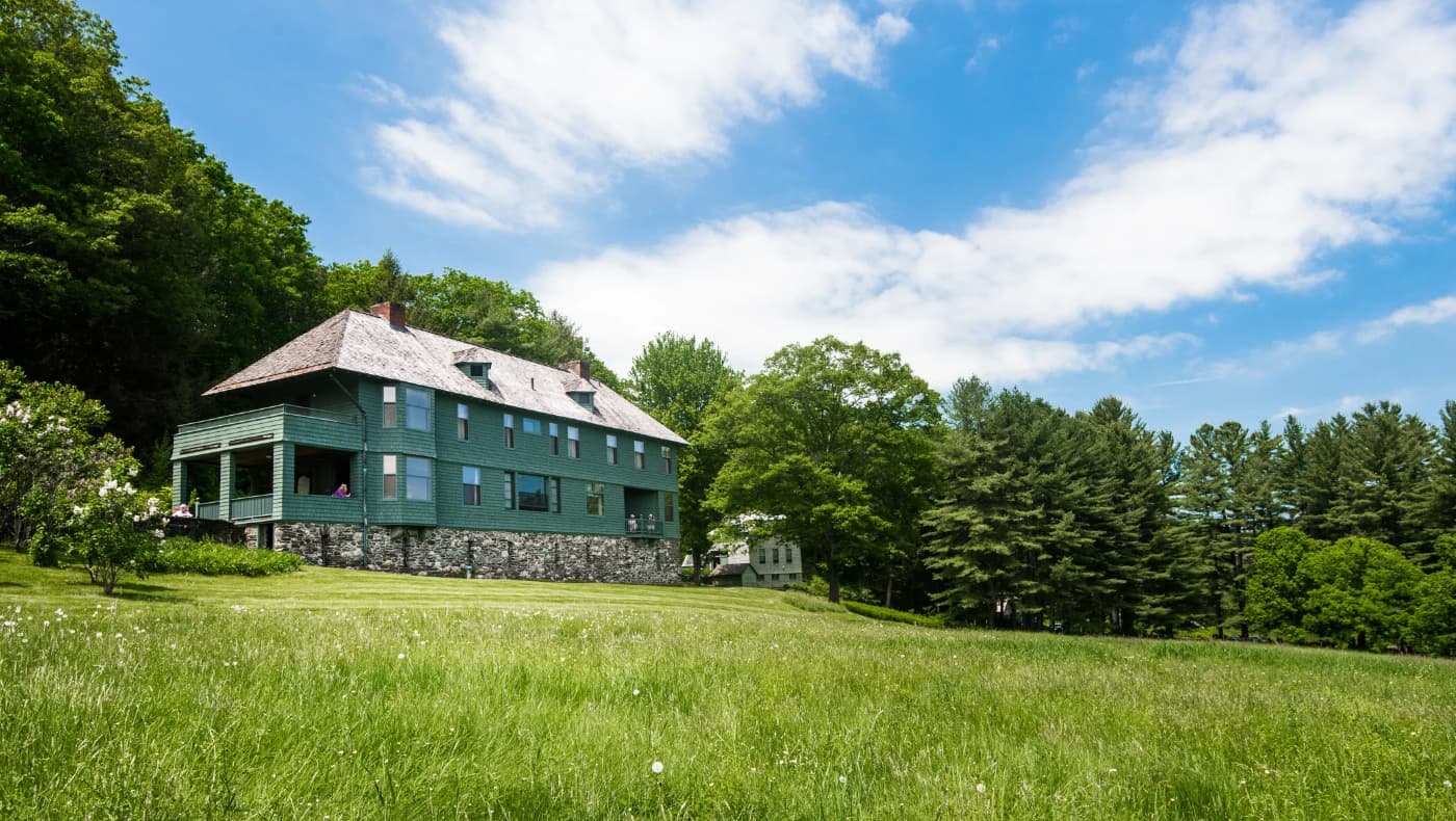 Exterior of Naulakha, Rudyard Kipling’s historic c. 1892 shingle-style home in Dummerston, Vermont. The elongated, ship-like house features dark wood shingles, a long covered porch, and stone foundations, nestled in a lush, wooded landscape.