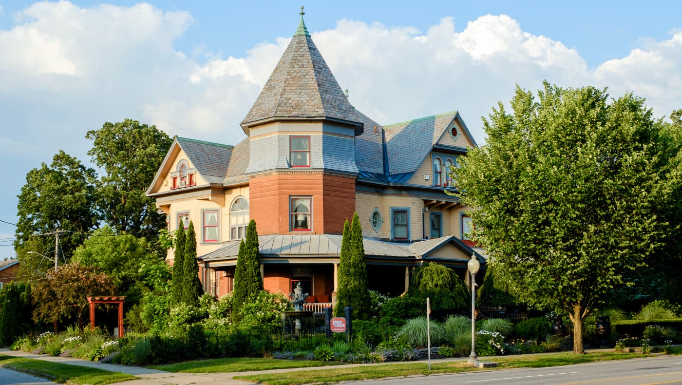Exterior of the Union Gables Inn, a stunning 1901 Queen Anne Victorian mansion in Saratoga Springs. The three-story building features a deep-red shingle and wood facade, a prominent corner turret with a conical roof, and a sweeping wraparound porch with white columns.