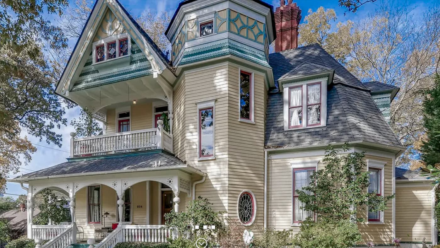 Exterior view of the Sugar Magnolia Bed and Breakfast, a grand 1892 Queen Anne Victorian home in Atlanta’s Inman Park historic district. The light-colored house features a prominent three-story corner turret, a wide wraparound front porch with classic columns, and intricate gables.