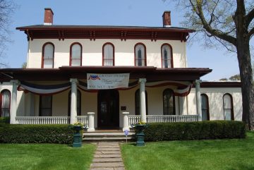 The front exterior of Ruthmere Mansion in Elkhart, Indiana. A three-story Beaux-Arts limestone and buff-brick residence featuring a grand symmetrical facade, a balustraded piazza, and a prominent porte-cochère.