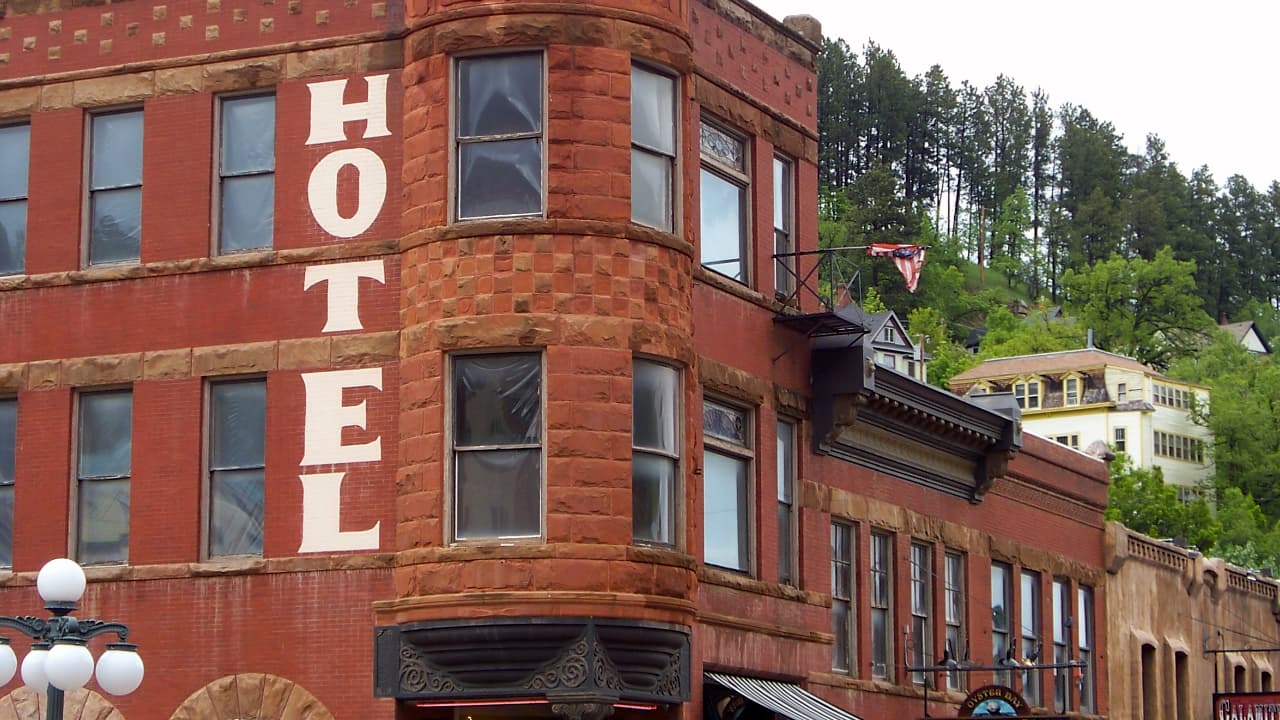 Exterior of the historic 1895 Fairmont Hotel in Deadwood, SD, featuring a rounded corner turret of rugged red sandstone, large vertical 'HOTEL' signage, and decorative wrought-iron window details.