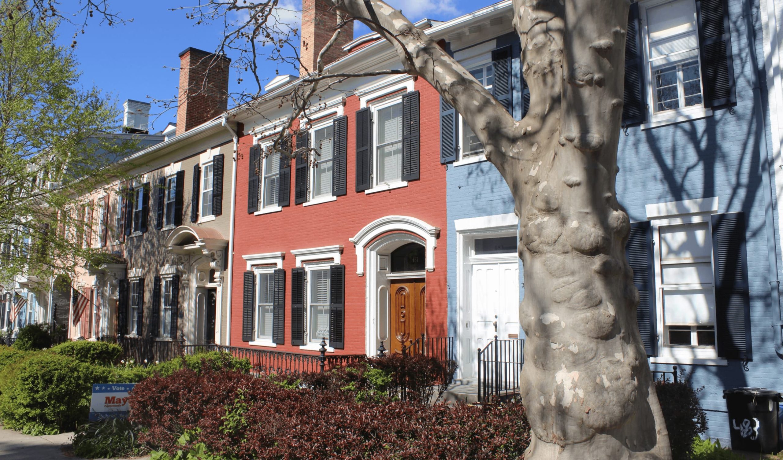 Exterior view of a historic Federal-style brick rowhouse on South Main Street in Geneva, New York. Part of the South Main Street Historic District, the building features three stories of well-preserved red brickwork, tall symmetrical windows with white-painted shutters, and a classic raised stoop entrance. The row is set against a bluff overlooking Seneca Lake.