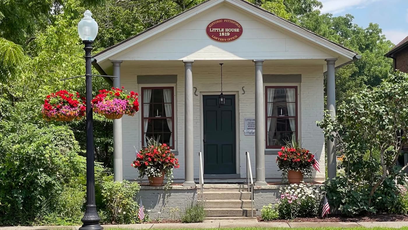 Exterior of the "Little Museum" in Pittsford, NY, a charming one-story historic brick building. The structure features a gabled roof, tall arched windows with white-painted shutters, and a simple wooden door.