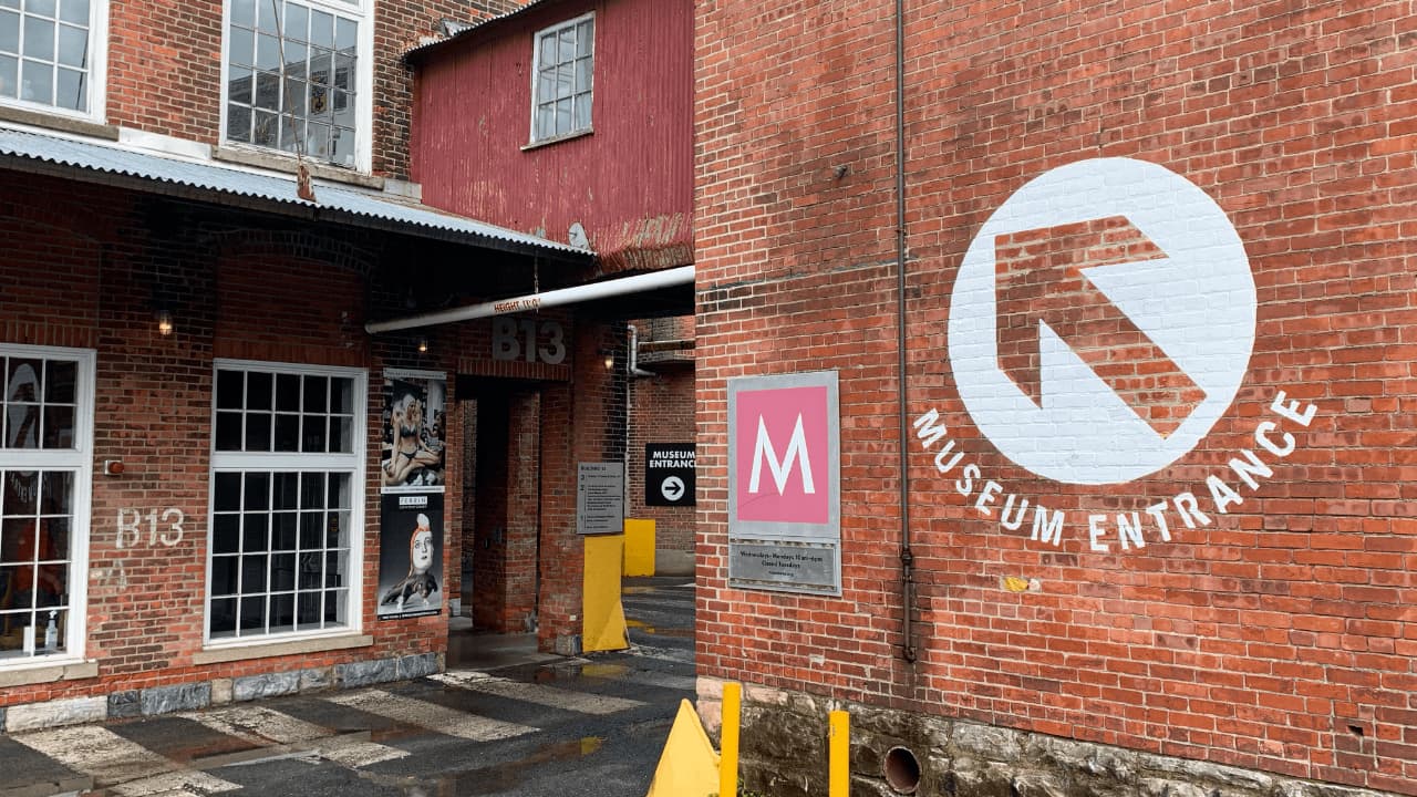 The main entrance to MASS MoCA in North Adams, MA, showcasing the adaptive reuse of a 19th-century brick textile mill complex with its iconic elevated 'bridge' walkway connecting historic industrial buildings.