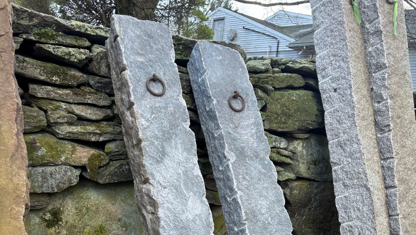 Two antique hand-split granite hitching posts with hand-forged iron rings leaning against a rustic stone wall.