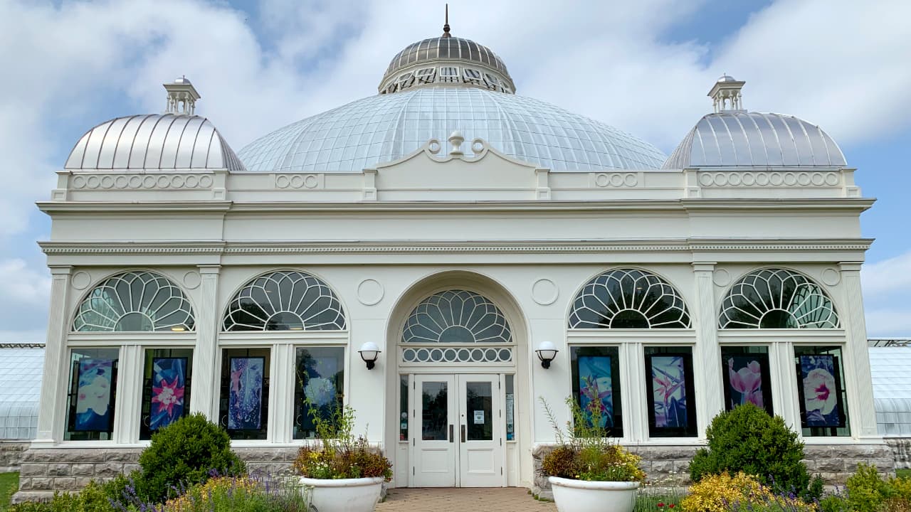 Exterior view of the historic Buffalo & Erie County Botanical Gardens conservatory in Buffalo, NY, featuring its iconic glass tri-dome design and Beaux-Arts architectural details.