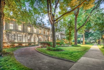 The Louis Kamper Mansion in Indian Village, Detroit. This 1915 Neo-Renaissance estate features a grand limestone facade, a symmetrical entry with a copper-roofed portico, and elegant arched windows surrounded by professional landscaping and wrought iron fencing.