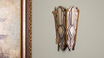 Vintage Art Deco wall sconce with a geometric white and cream stained glass shade, mounted on a sage green wall next to an ornate gold picture frame.