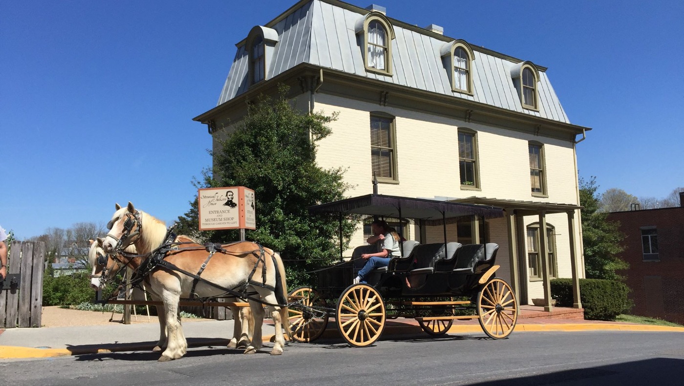 Two blond draft horses pulling an open carriage past a historic building with a metal mansard roof in Lexington, Virginia.