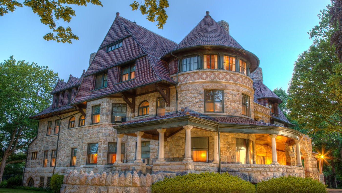The Oliver Mansion in South Bend, Indiana, a Queen Anne fieldstone mansion with a round tower and wide porch, lit up at dusk.